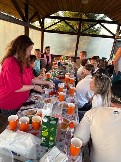 The image shows a group of people gathered around a long table covered with food and drinks in what appears to be an outdoor covered structure, possibly a gazebo or pavilion. The individuals on one side of the table are leaning in, seemingly engaged in eating or looking at the food. The people on the other side of the table are seated and interacting. The setting appears to be a casual gathering with orange cups, bottled water, juice boxes, and plates of food scattered across the table. The structure has wooden supports and transparent walls, revealing trees and foliage outside. The location might be in Кузнецово, Russia, although specific landmarks are not visible in the image to confirm this.