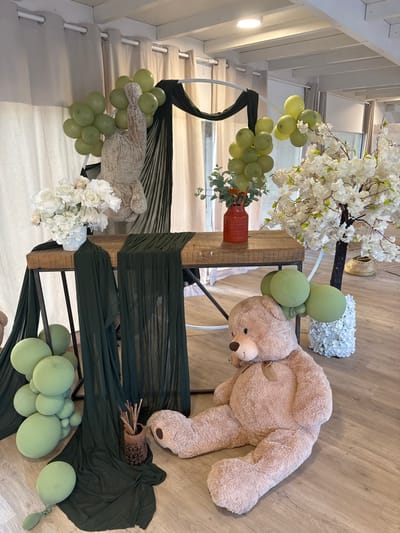 The image depicts a party or event setup with a rustic, natural theme. The scene is indoors, likely in a venue in Hem, France, characterized by a wooden floor, exposed ceiling beams, and neutral-colored curtains.
Dominating the foreground is a very large, light brown teddy bear, seated on the floor, facing to the left. Beside the teddy bear, to its right, is a decorative tree with white blossoms. In the mid-ground, a rustic wooden table with dark metal legs is set up. A dark green fabric runner drapes across the table, and more of the same fabric is draped around the table legs and the general area.
On the table, there is a white vase filled with white flowers, some of which appear to be roses and hydrangeas. To the right of the flower arrangement, a red vase holds greenery and some orange decorative elements.
Numerous light green balloons are scattered throughout the scene. A large cluster of balloons is positioned to the left of the table, extending upwards. Smaller clusters of balloons are also visible near the teddy bear and behind the table. In the background, a large, circular metal arch is partially visible, draped with black and dark green fabric. A second, smaller teddy bear or stuffed animal is positioned behind the table, with only its upper body and arms visible, holding a bunch of balloons.
The overall impression is a carefully arranged decorative display for an event.