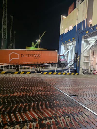 The image shows a night scene at the port of Ravenna, Italy. A large ferry, painted with blue and white, is docked. On the deck of the ferry, an orange truck trailer with the logo "Di Stefano trasporti" is visible, along with a red tow tractor. The deck of the ferry is made of metal with a distinctive chevron pattern, appearing wet and worn. In the background, tall cranes stand against the dark night sky, illuminated by lights. The overall impression is one of industrial activity at a port during the night.