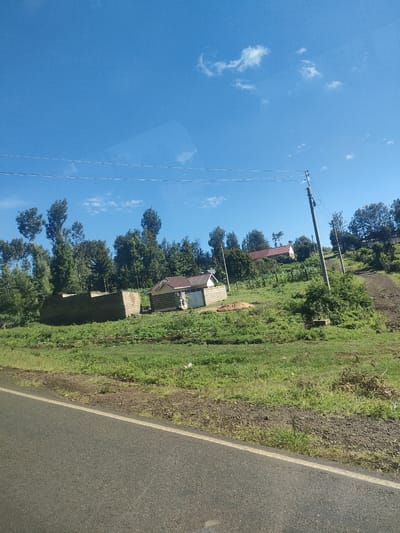 The image shows a rural landscape in Kenya, likely within a municipality ward. The sky is blue with scattered clouds. In the foreground, there's an asphalt road with a white painted line marking the edge. Beyond the road, a grassy hillside rises, dotted with trees and several buildings. One building is partially constructed, appearing as a low brick wall structure without a roof. Another building is more complete, with walls and a reddish tile roof. A third structure is visible in the distance with a red roof. Power lines run across the scene. A utility pole stands on the hillside. A dirt path leads up the hill. The environment is green and appears to be sparsely populated.