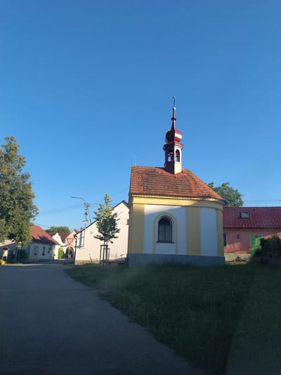 This is an eye-level, outdoor, full shot of a small village street in Bošovice, Czechia, on a clear, sunny day. The sky is a vibrant blue with no clouds. In the foreground, a paved road curves to the left. To the right of the road is a patch of green grass with some weeds. In the middle ground, a yellow and white building with a red tiled roof and a small bell tower stands out. Adjacent to it, to the right, is a pink building with a red tiled roof and a green door. Behind these buildings, more houses with red roofs are visible. A tall green tree is on the left side of the image, and a smaller tree is in front of the yellow and white building. A street lamp is visible behind the smaller tree. The scene is peaceful and quiet, with no people or animals visible.