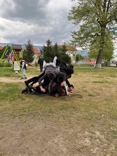 A group of approximately 10 young people are engaged in a physical altercation on a grassy area in Karavelovo, Bulgaria. Some are standing, while others are piled on top of each other on the ground. A playground swing set is visible in the foreground. A soccer ball is near the swing set. Buildings and trees surround the grassy area. The sky is overcast.