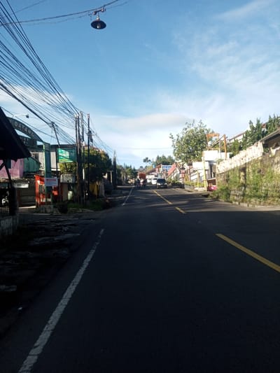 A daytime street scene is observed, identified by the user as being in the city of Khouathineung, Laos. However, prominent commercial signage includes terms such as 'SELAMAT DATANG,' 'ANEKA PULSA,' and 'PELAJARAN,' which are in Bahasa Indonesia or Bahasa Melayu.
The asphalt road, marked with a solid white line on the left edge and intermittent yellow center lines, curves gently uphill and to the right into the distance. Long, distinct shadows extend from the left (viewer's perspective) across the road, indicating a low sun angle, likely morning.
On the left side of the road, an unpaved, uneven shoulder borders a series of utility poles supporting a dense tangle of overhead electrical and communication wires. A single, dark-colored street light fixture hangs suspended from these wires high above the road. Behind the poles, various low-rise commercial or residential structures display faded paint (e.g., pink, green) and host multiple signs with text in the aforementioned languages, including "CANTEEN."
The right side of the road features a stone retaining wall, partially covered with moss and vegetation, supporting an elevated residential area with various buildings and dense foliage, including slender trees resembling conifers.
In the mid-ground, a white multi-purpose vehicle (MPV) travels away from the viewer on the right side of the road, accompanied by several indistinct motorcycles. No identifiable individuals are clearly visible. The background reveals additional buildings, vegetation, and distant hills under a mostly clear blue sky with wispy white clouds.