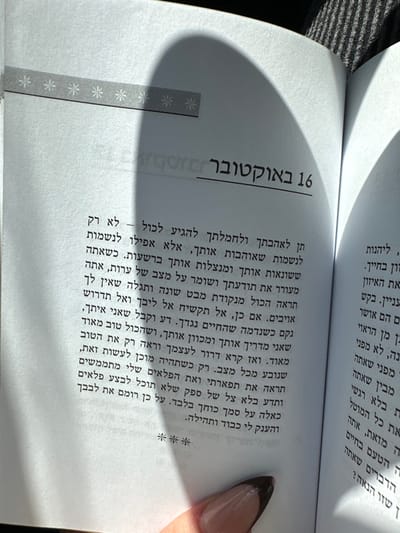 The image displays a close-up, top-down view of an open book held by a person's hand in Kafr Kasim, Israel. The book's pages are white, with the left page prominently featuring a heading in Hebrew that translates to "16 October," followed by columns of Hebrew text. A grey decorative strip with white asterisk patterns is present across the top left edge of the left page. The right page also contains visible Hebrew text. A distinct, dark, curved shadow extends across the upper half and center of both pages, partially obscuring text and the decorative strip, indicating strong directional lighting from an unseen source. A human hand, with fingernails painted with dark tips, is visible at the bottom right corner, holding the book open.