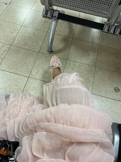 An individual, seen from a high angle, is seated in a waiting area, possibly an airport or hospital, with a light pink dress flowing around their legs and a matching pink sneaker visible on their right foot. The floor is tiled in a neutral beige color, and a metallic bench with a perforated seat is present in the background. The scene is indoors, suggested by the artificial lighting and the nature of the seating.
