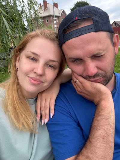 A selfie shows two people in an outdoor setting, likely a park or waterfront, with buildings and foliage in the background. The woman, with blonde hair and freckles, is on the left, smiling and leaning her head on the man's shoulder. She is wearing a light blue t-shirt and has long, light pink fingernails. The man, on the right, is wearing a blue t-shirt and a dark baseball cap. He has a beard and is winking, with his hand resting on his cheek. A gold wedding ring is visible on his left hand. The background includes a weeping willow tree, two-story buildings with red tile roofs, a fence, and a cloudy sky. The location appears to be the embankment in Breytovo, Russia, near the confluence of the Sit and Mologa rivers.