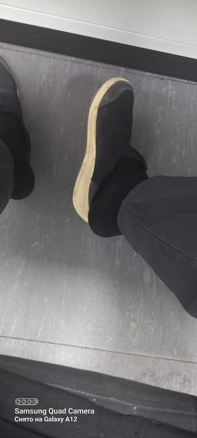 The image shows a low-angle shot of someone's legs and feet wearing black pants and black socks with slip-on sneakers. The sneakers have a light-colored sole and appear to be slightly worn. The person is standing on a grey, textured floor, possibly linoleum or tile, with a white vertical element to the left of the frame. A black vertical element, possibly a doorway or partition, is present between the white element and the floor. The lighting suggests an indoor setting. There is a watermark in the bottom right corner that reads "Samsung Quad Camera Снято на Galaxy A12" in white text. The activity appears to be simply standing. No specific time of day, weather conditions, or strong emotional cues are discernible. The location context provided is Levakant, Tajikistan.