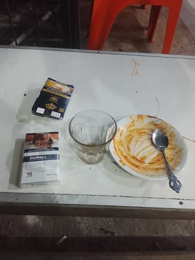 On a white, likely melamine, table sits a collection of items: two packs of cigarettes (one Dunhill Fine Cut Mild and one Maestro Edition), a mostly-empty glass, and a white plate smeared with the remnants of a meal and a silver spoon. The scene suggests a solitary individual finished eating and is about to smoke. An orange plastic chair is partially visible in the background. The table is situated outdoors, evidenced by the concrete ground below and the dimly lit area behind the table, with possible metal bars. The OCR indicates the location is Dayahlhoksukon Barat, Indonesia, due to the Indonesian language on the cigarette packs.