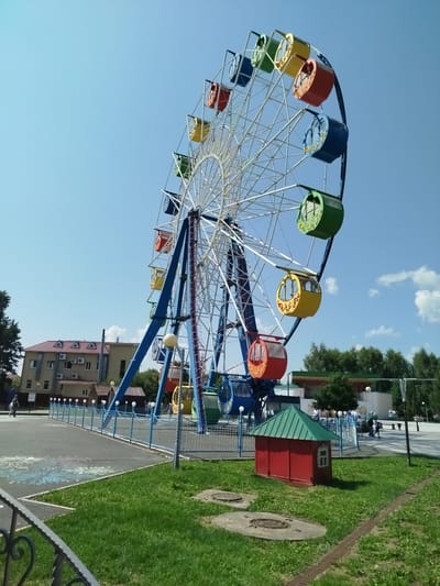 The image shows a Ferris wheel in an amusement park located in Yalutorovsk, Russia. The Ferris wheel is tall and colorful, with red, yellow, green, and blue cabins. It is situated on a clear blue day with a few white clouds in the sky. In the background, there is a building with a red roof and several air conditioning units. A small red building with a green roof is in the foreground, and there is a metal fence surrounding the park. A few people are visible in the background, walking around the park. The ground is a mix of asphalt and grass.