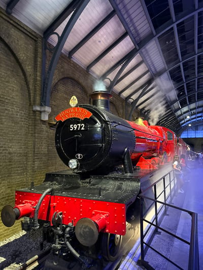 The image shows the Hogwarts Express steam locomotive, numbered 5972, at Platform 9 ¾ in King's Cross Station, London, United Kingdom. The locomotive is red and black, with steam emanating from its chimney. The platform is lined with a black metal railing and a brick wall. Several people are visible on the platform, including tourists posing for photos. The station's arched ceiling is visible in the background. The scene is well-lit, with artificial lighting casting a blueish glow on the platform.