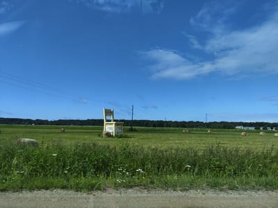 The image depicts a rural landscape under a bright blue sky with scattered white clouds. In the foreground, a gravel road is visible, with a grassy field extending beyond it. The field is dotted with numerous round hay bales. A prominent feature in the field is a large, chair-shaped wooden structure with a sign that reads "RAKARI." Below this structure, there is a smaller, white building with signs that say "ATVERTS OPEN." In the background, a dense line of dark green trees forms the horizon, with a few buildings and power lines visible in the distance. The scene appears to be a roadside attraction or farm stand in Svētciems, Latvia.