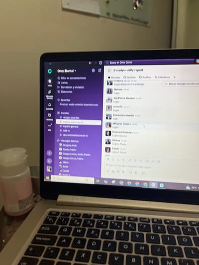 An eye-level, tilted shot captures a laptop displaying a communication platform, likely in an indoor setting in San Luis, Peru. The laptop screen is the central focus, showcasing what appears to be a Slack-like application with a purple sidebar and a white main chat window.
On the screen, the application is branded "Omni Dental." The left sidebar organizes content under categories like "Hilos de conversaciones" (Conversation threads), "Juntas" (Meetings), "Borradores y enviados" (Drafts and sent), and "Directorios." Several channels are listed, including "#design-pixel-lab," "#equipo-daily-report" (team daily report), "#equipo-general," and "#ops-automatizaciones-la." Direct messages to various individuals, such as Gregory Arcia, Cande, Mario, and Javier, are also visible.
The main chat window is focused on the "#equipo-daily-report" channel, showing a series of messages indicating team members' activity. Entries like "Stefany 04:30 Login," "Van Pierre Jimenez 04:39 Login," "Patrick Nacimento 03:02 Login," and "Milagros Gomez 03:38 Login" are visible, along with "Logout pausa" entries for Federico Gonzalez (07:07), Silvana (07:08), and Cande (07:08). A search bar at the top right reads "Buscar mensajes en este canal," and a small weather icon indicates 22°C.
The right side of the image reveals part of the laptop's physical keyboard, which features a Spanish QWERTY layout with keys like 'Ñ' and "bloq mayús" (caps lock). In the immediate foreground, a light pink container or bottle is partially visible. The background shows a plain, light-colored wall. The overall scene suggests a work environment where team members communicate and track daily activities.