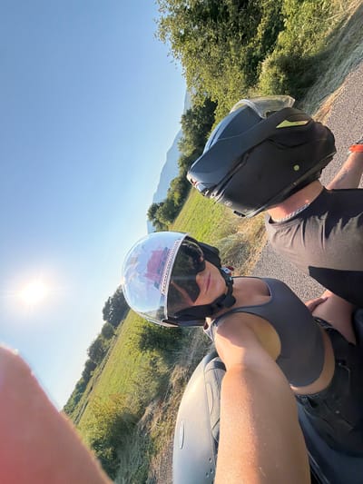 Two people, wearing motorcycle helmets and casual clothing, are taking a selfie on a motorcycle. The person in the foreground is wearing a black tank top and sunglasses, while the person behind them is wearing a black t-shirt. Both are wearing black helmets. The scene is outdoors, with a grassy field and trees visible in the background, under a clear blue sky with a bright sun. The motorcycle appears to be parked on the side of a road. The time of day is likely late afternoon, given the angle of the sun. The overall mood seems casual and relaxed. The text "REB38" is visible on the side of one of the helmets. The location context is Buljarica, Montenegro.