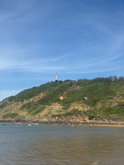 The image shows a coastline with a steep, vegetated hill on the left and the ocean on the right. A narrow beach with large rocks lines the base of the hill. A lighthouse is visible on top of the hill, surrounded by trees and vegetation. Several people are scattered along the beach and in the shallow water. One person appears to be swimming or wading. The sky is clear and blue with a few wispy clouds. The water is a murky green color. The scene appears to be set in a tropical or subtropical location, likely during daylight hours given the clear sky. The overall mood is peaceful and leisurely, suggesting a day at the beach. There is no visible text in the image.