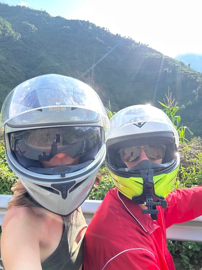 Two individuals wearing full-face motorcycle helmets are depicted in a close-up selfie taken on a sunny day in the mountainous region of Tú Lệ, Vietnam. The person on the left wears a silver "Royal" branded helmet with a reflective visor that shows a sharp curve in a paved road and the reflection of an arm holding a smartphone used to capture the image. This individual has visible light brown hair and wears a dark sleeveless top. The person on the right wears a white and neon yellow helmet with an attached GoPro chin mount, sunglasses visible under the raised visor, and a red jacket with white trim. Both subjects are positioned alongside a metal guardrail, with dense green vegetation, including corn stalks, visible in the foreground and a vast, verdant mountain range under a clear sky in the background. Sun flares are prominent, indicating direct sunlight from the upper right.