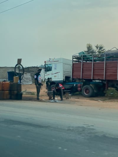 A large truck with a red cargo container on the back is parked beside a road. Two people work on the truck's tire. A man wearing what appears to be a uniform and holding a rifle stands guard near the truck. A stack of barrels and a metal hoop are near the guard. A small building is beside the road.