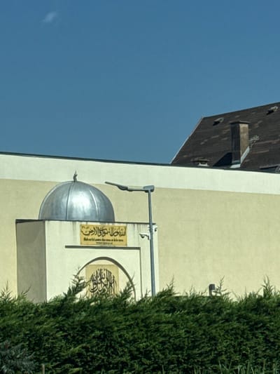 The photograph depicts the exterior of a building, likely a mosque, with a small dome visible. A rectangular sign with Arabic text and French translation is mounted on the wall. A larger, decorative panel with Arabic calligraphy in an arched frame is also present. A portion of a roof is seen in the upper-left. A dense green bush partially obscures the building's facade on the right. The sky is a clear, bright blue. The building appears to be cream or light yellow in color.