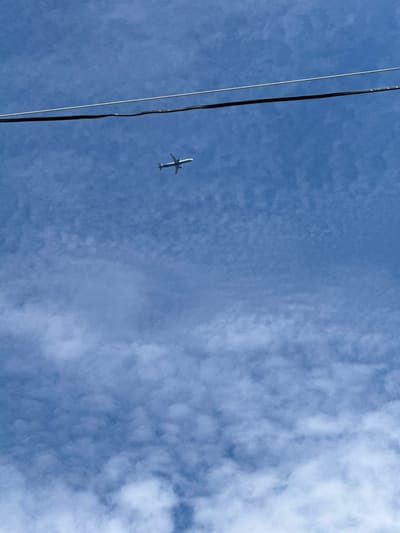 The image shows a clear blue sky with scattered cumulus clouds. Two wires are visible horizontally across the upper portion of the frame, likely utility or power lines. A white airplane is flying in the distance against the blue sky. The overall scene is natural and likely captured outdoors. Without further information or context, it is impossible to determine the specific location within Дзержинец-83 Снт, Russia.