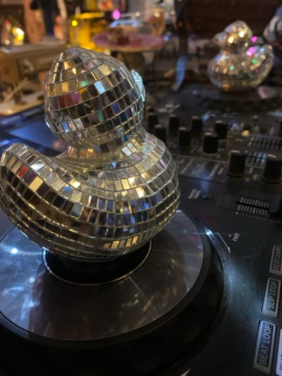 A close-up shot displays a silver, disco-ball-shaped object, attached to a black DJ turntable. The turntable has various buttons and sliders visible, including "BEAT JUMP", "SLIP LOOP", and "BEAT LOOP" labels. A second, blurred disco ball is visible in the background, suggesting an indoor setting, possibly a bar or entertainment venue in Baan Nai Suan, Thailand. The lighting is dim with reflective surfaces, enhancing the disco theme.