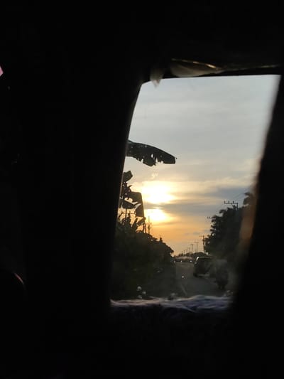 The photo, taken from within a vehicle in Kruengseupeng, Indonesia, shows a road stretching into the distance, framed by what appears to be a partially obscured window or opening. The sun is setting, casting a warm, orange glow over the scene and silhouetting the roadside trees and vegetation, which include banana trees. Utility poles line the road. In the distance, vehicles are visible. The immediate foreground inside the vehicle is dark and out of focus.