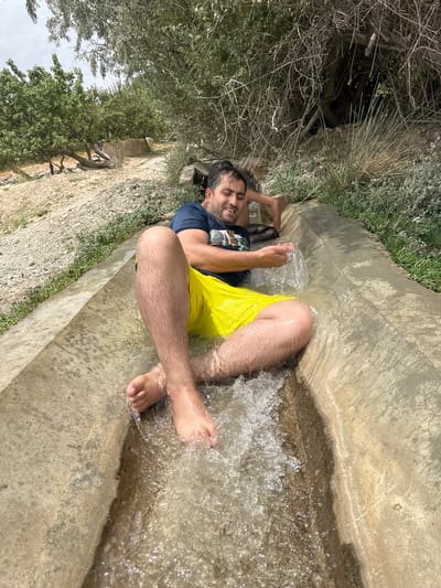 A man is lying on his back in a concrete irrigation channel filled with water, located in Kan Mehtarzai, Pakistan. He is wearing a dark blue t-shirt and bright yellow shorts. His arms are bent, with his right hand splashing water. His legs are extended, with his feet submerged in the flowing water. The channel is surrounded by dry, rocky terrain and sparse vegetation, with trees visible in the background under a cloudy sky.