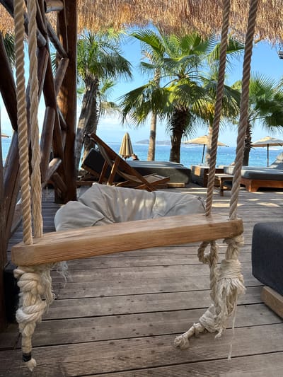 The image depicts a wooden swing with thick ropes attached, hanging over a wooden deck. In the background, palm trees frame a clear blue sky and the sea, suggesting a tropical or Mediterranean coastal location. The deck has several lounge chairs and umbrellas, indicating a beach resort or outdoor seating area. One can infer this scene is likely located on the island of Thassos, Greece, given the characteristic landscape and architecture often found in such areas. The lighting suggests it is daytime.