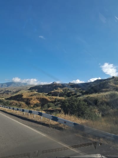 The image depicts a scenic view of a mountainous landscape under a clear blue sky. A highway guardrail runs along the side of a paved road in the foreground, separating it from a dry, grassy hillside dotted with sparse green bushes. In the distance, rugged mountains rise, with some areas appearing hazy. The overall impression is one of a rural, possibly arid, environment. The location is identified as the city of Vayk, Armenia.