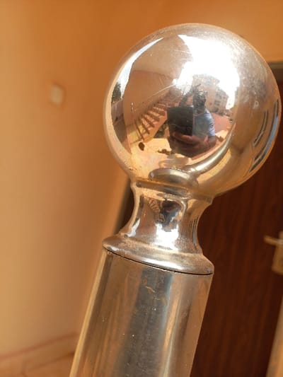 A close-up, angled photograph captures a reflective spherical metallic finial atop a cylindrical metallic post, likely part of a railing, located in Oturkpo, Nigeria. The polished metallic surface of the finial exhibits minor scratches and specular highlights. The finial's reflection prominently displays a male individual, presumably the photographer, holding a dark-colored smartphone or camera. This individual wears a light-colored, possibly striped short-sleeved shirt and has short dark hair or a beard. Behind the photographer, the reflection reveals an outdoor scene featuring a multi-level staircase with visible railings, several light-colored multi-story buildings, and sparse green vegetation under a bright sky. The immediate background to the left of the finial shows a blurred, plain light-orange wall with a faint rectangular lighter patch. To the right, a blurred dark brown wooden door with a metallic handle is partially visible. The overall scene is illuminated by natural light, enhancing the reflective properties of the metal.