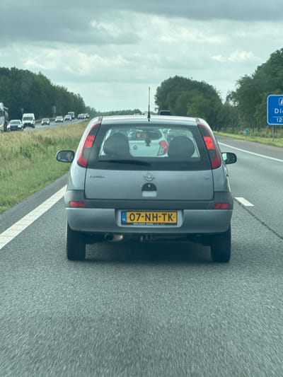 A gray Opel Corsa hatchback, license plate "07-NH-TK", is driving on a highway in the Netherlands. The car's brake lights are illuminated. The road is asphalt and has white lane markings. Trees and a grassy verge are visible on the left side of the road. On the right side, a blue highway sign indicates "Diedrichsland" and "12 km". The sky is overcast and cloudy. The image is taken from behind the car, slightly to the left.