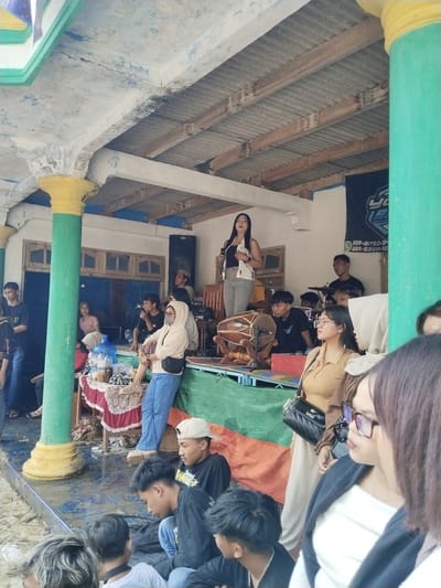 The image shows a group of people in Jenawi, Indonesia. A woman is standing on a raised platform, speaking into a microphone. Behind her, a band is set up with drums and other instruments. Several people are sitting or standing around the platform, watching the performance. The setting appears to be an outdoor event, possibly in a covered area with pillars. The pillars are painted with yellow and green bands. The floor is wet.