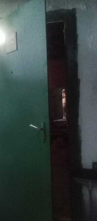 The image depicts an interior scene, likely a hallway or doorway, with a green door dominating the left side of the frame. The door has a silver handle and a keyhole. To the left of the door, a small, light-colored plaque is mounted on the wall, featuring some text which is not clearly legible. The right side of the image shows a dark opening, possibly another doorway or a narrow passage, leading into a dimly lit space. The lighting suggests it might be evening or a poorly lit interior. The walls appear to be painted in muted tones, with some texture visible. The overall impression is one of a functional, perhaps somewhat utilitarian, indoor environment. There are no people visible in the image.