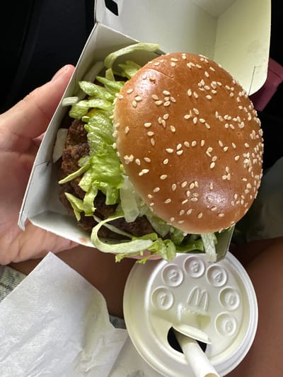 A person is holding a McDonald's Big Mac in its cardboard container and a McDonald's soda cup with a straw in a car. A white napkin is next to the cup. The Big Mac features a sesame seed bun and lettuce. The location cannot be determined from the image.