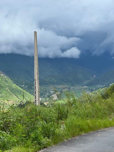 An outdoor, eye-level shot captures a tall, weathered concrete pole standing vertically in the foreground, positioned to the left of center. The pole is segmented with rectangular openings, and holes are visible along its length. It stands amidst lush green foliage, which covers the lower portion of the frame. To the right, a grey asphalt road curves away from the viewer.
The mid-ground reveals a picturesque valley with rolling green hills and mountains that recede into the distance. A small village with scattered buildings and agricultural fields is visible within the valley. A river or stream meanders through the landscape.
The background is dominated by a dramatic sky with dark, heavy clouds, suggesting an overcast or stormy day. The lighting is diffused, casting a soft light over the entire scene. The image appears to be taken in Lùng Tám Commune, Vietnam, given the characteristic mountainous terrain and vegetation. No people or specific actions are depicted.