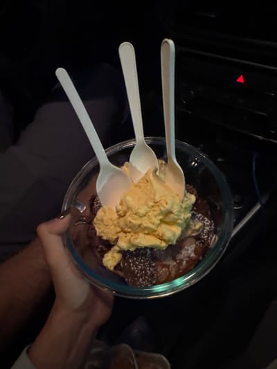 A clear bowl of dessert, likely brownie or cake, is topped with a large scoop of yellowish ice cream and powdered sugar. Three white plastic spoons are placed haphazardly on top. The bowl is held by a hand with a ring on the ring finger. The scene appears to be inside a car at night, given the dashboard and dim lighting. Another container sits in the background on the right.