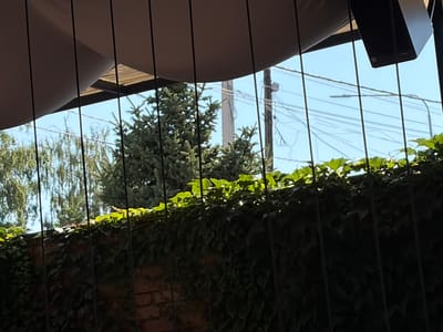 The image shows a view through vertical cables, possibly part of a fence or structure, looking towards trees and a brick wall covered in ivy. The sky is blue with some utility lines and poles visible. In the upper part of the image, a light-colored canopy or awning is present. The scene appears to be outdoors in a somewhat overgrown urban or suburban environment. It is not possible to determine the specific location in Çolpon-Ata, Kyrgyzstan, from the visual information provided.