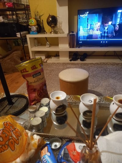 A room scene features a television atop a white shelf, displaying a program. Beside it sits a tan drum. A mirrored tray laden with teacups on sticks, candles, and packaged snacks dominates the scene. Underneath, a bag of Alesto roasted peanuts is visible. A black fan stands in the foreground, partially obscuring a bag of flavored puffed corn snacks. A carpet with a gray pattern spreads across the floor.