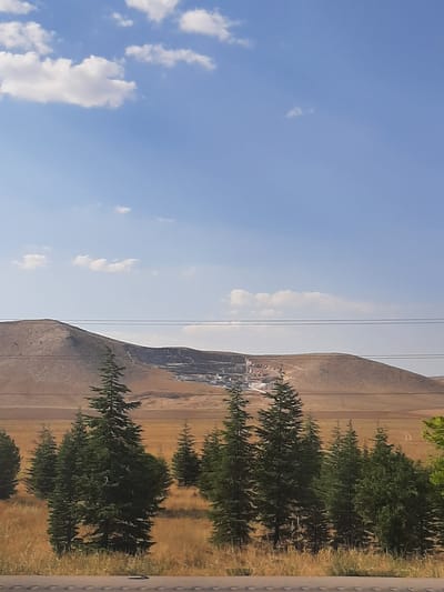 A wide shot captures a vast, arid landscape under a bright blue sky dotted with fluffy white clouds. The foreground is dominated by a line of evergreen trees, their dark green foliage contrasting with the dry, golden-brown grasses and scattered bushes behind them. In the mid-ground, a large, excavated quarry is visible on the side of a sloping hill. The quarry's white and light-colored rock faces appear to be artificially carved, creating tiered levels and revealing geological strata. Power lines stretch horizontally across the scene, suspended between poles that are not fully visible. The distant background consists of rolling, barren hills under the expansive sky. The scene appears to be a natural environment impacted by human activity, likely an open-pit mine in the Karatay district of Turkey. There are no people or animals visible in the image.