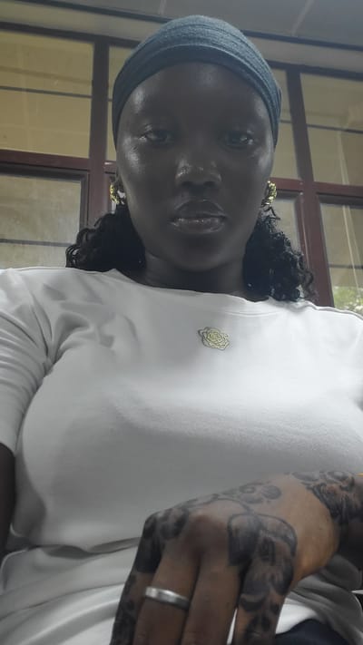 A medium shot captures a Black woman with dark skin, wearing a dark blue headscarf and a white t-shirt with a gold rose-shaped brooch. She is positioned in what appears to be an indoor setting, possibly an office or classroom, indicated by the presence of large windows with wooden frames in the background. The woman's face is directed towards the camera, with her lips slightly parted and her eyes looking straight ahead. She is wearing gold hoop earrings, and her hair is visible as dark curls cascading down. Her left hand, adorned with intricate dark henna tattoos and a silver ring, rests on her chest, obscuring a portion of her t-shirt. The lighting in the image is somewhat dim, casting subtle shadows on her face and highlighting the sheen on her lips. The overall impression is a candid portrait taken indoors.