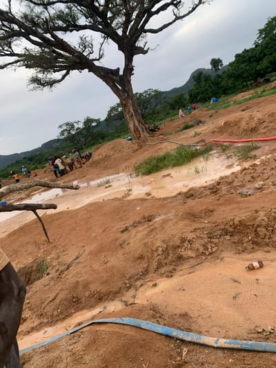 The image depicts a muddy, excavated hillside in Buga da Gindi, Nigeria. Several individuals are engaged in what appears to be mining or earth-moving activities, with some digging and others carrying materials. They are scattered across the slope, with groups forming around areas of activity. The landscape is dominated by reddish-brown soil and large puddles of murky water, indicating recent rainfall or water usage for excavation. Sparse vegetation, including a large tree in the foreground and trees on the distant hills, dots the scene. Hoses, one red and one blue, are visible, suggesting the use of water for the process. There are also discarded bottles and debris present on the ground. The sky is overcast, contributing to a diffused lighting.