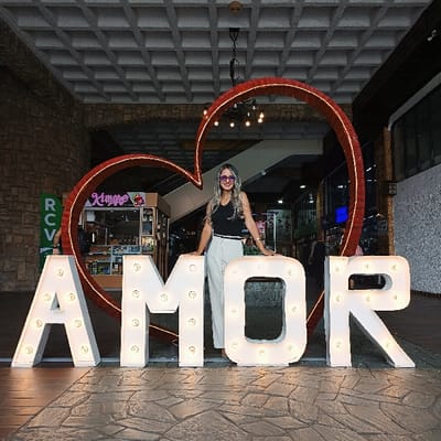 A woman stands in front of a large "AMOR" (love in Spanish/Portuguese) sign, each letter illuminated with bulbs. The sign is placed beneath a large red heart made of lights, creating a romantic backdrop. The woman has on sunglasses, a black top and light pants. She stands in what appears to be an indoor commercial space with shops and stone facades in the background. The scene is well-lit, emphasizing the glow of the signs.