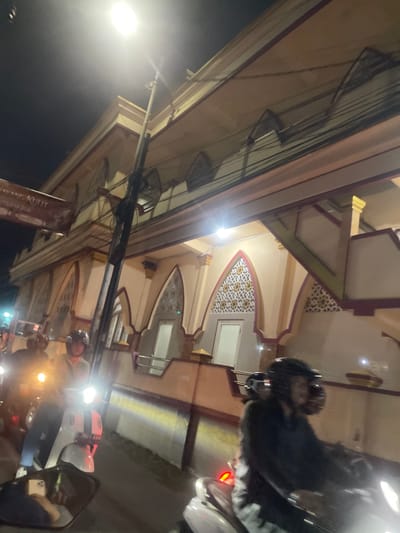 This image captures a street scene at night in Sawotratap, Indonesia. The primary focus is on a building with distinct architectural elements, featuring arched openings decorated with intricate, geometric patterns in yellow and white against a muted background. The building's facade is adorned with maroon and cream colored trim. Above the building, a complex network of electrical wires is visible, strung between poles.
In the foreground, several individuals on motorcycles are present, suggesting a street or thoroughfare. One person, wearing a helmet, is seated on a motorcycle on the right side of the frame, partially obscured by glare. Another motorcycle, with a person wearing a helmet and dark clothing, is also visible. A bright, white light from a streetlamp illuminates the left side of the image, casting a strong glow and contributing to the nighttime atmosphere. The overall impression is of a bustling urban environment at night, with the architectural beauty of the building contrasting with the utilitarian network of wires and the presence of motorbikes. No specific event or emotion is clearly conveyed, but the nighttime setting and the presence of vehicles suggest general urban activity. A portion of a sign with text is visible in the lower left, but the text is illegible.