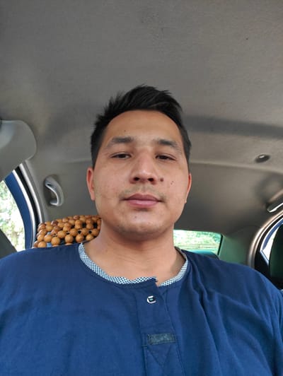 A close-up, waist-up, selfie-style photograph shows a man sitting in the driver's seat of a car. The man has dark hair, brown eyes, and is looking directly at the camera. He is wearing a dark blue shirt with a checkered collar and two buttons. Behind him, a string of wooden beads is visible, likely a car accessory for the seat. The interior of the car, including the headliner and a handle, is visible. Through the car windows, greenery and trees can be seen, suggesting an outdoor setting, possibly on a street in Navoiy City, Uzbekistan. The lighting suggests it is daytime.