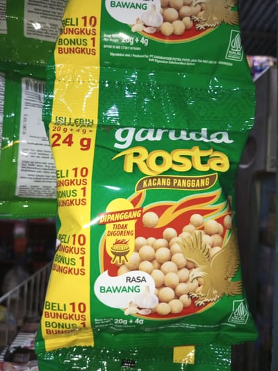 The image shows a close-up of a green plastic bag of Garuda Rosta Kacang Panggang (roasted peanuts). The bag has a yellow vertical stripe on the left side with red text that reads "BELI 10 BUNGKUS BONUS 1 BUNGKUS" (Buy 10 packs, get 1 pack free). Above this, also on the yellow stripe, is text that translates to "GET MORE FREE" and "20g + 4g = 24g". The main part of the bag features the brand name "Garuda Rosta" in large yellow letters, with "KACANG PANGGANG" written below in smaller brown letters. A graphic of an eagle is depicted on the right side of the bag, with a pile of roasted peanuts in front of it. There is a white circle with an image of a garlic bulb and the text "RASA BAWANG" (onion flavor). The top of the bag shows a graphic of garlic cloves and a peanut. A "HALAL" logo is visible on the bottom right. The background is blurred and appears to be a retail environment with other packaged goods visible. The location is not specified in the image, but the text "INDONESIA" is visible on the bag, indicating the country of origin.