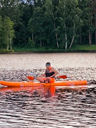 A young person wearing goggles and a life vest sits on an orange stand-up paddleboard on a lake. The paddleboard is labeled "AQUA MARINA" and "FUSION". The person is holding an orange paddle. The lake's surface is textured with light and dark patterns. In the background, a dense forest with a variety of trees lines the shore of the lake. The sky is not visible. The scene appears to be in a natural outdoor environment.