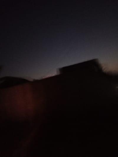 The image captures a blurry, low-light scene, likely taken during dusk or twilight. The upper portion of the frame displays a dark, gradient sky, transitioning from a deeper blue-grey at the top to a slightly lighter hue towards the horizon, with a subtle hint of orange-brown on the far right.

The lower half of the image is dominated by very dark, indistinct shapes. On the right side, a prominent rectangular silhouette, heavily blurred, appears against the lighter part of the horizon, suggesting a building or a large structure. The foreground and midground on the left and bottom consist of amorphous, dark masses, with some areas revealing a reddish-brown tint, possibly indicating obscured landforms or objects.

The overall blurriness and lack of sharp detail throughout the image suggest significant camera shake or motion blur, indicative of a long exposure in dim lighting conditions, or a photograph taken from a moving vehicle. No specific people or identifiable objects are clearly discernible. There is no visible text. The image does not contain specific visual cues to definitively identify Mururi, Kenya, beyond the provided context.