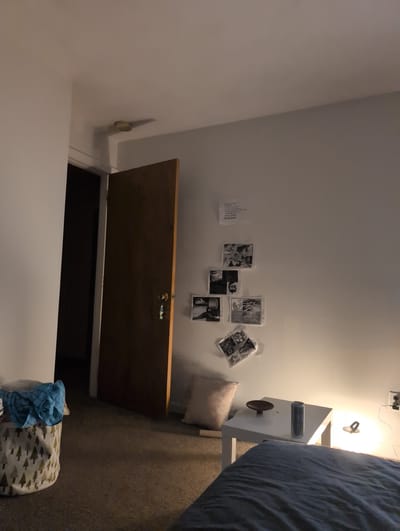 This image depicts a cozy, lived-in room, likely a bedroom or personal living space, with the perspective looking downwards towards a floor hatch. On the right side of the frame, a bed is visible, covered with a blue-grey duvet. Adjacent to the bed is a white bedside table, holding a dark cylindrical object (possibly a water bottle), a shallow brown bowl, and a lamp that emits a warm, bright glow, suggesting it is on and illuminating the area.

The left wall is light-colored and adorned with several small, black-and-white photographs or prints, taped or tacked onto the surface. Visible text on these prints includes "BEST FRIEND JOKES" and "OPEN." Further to the left, a document or paper with Cyrillic text, including the word "ВИЖДАМ," is also attached to the wall. High on this wall, an electrical outlet with wires is visible.

A prominent feature in the foreground is an open wooden hatch or door in the floor, revealing a dark, recessed space below, possibly leading to another level via stairs or a ladder. The hatch features a brass-colored latch mechanism. The floor around the hatch appears to be covered with a dark carpet or rug. In the lower-left corner, a fabric storage basket with a pattern of small green trees contains crumpled blue plaid fabric.

The room appears to be a private residence in Waltham, United States of America. The primary light source is the bedside lamp, indicating that the photograph was likely taken in the evening or at night. The scene is static, devoid of people or activity, focusing on the personal elements and architecture of the space.