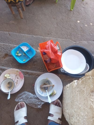 In Kimamba, Tanzania, the scene is an outdoor ground-level area with remnants of a recently consumed meal. At the bottom center, a person's feet are visible, clad in white, open-toed sandals and blue trousers. To their left, a round, light-colored plate with floral patterns holds a white bowl containing a silver spoon, and several lime wedges and food scraps are on the plate. To their right, another round, white ribbed plate supports a white bowl with a blue pattern, containing a bone fragment, a lime wedge, and a silver spoon. A crumpled clear plastic bag lies between these plates.
In the upper left, a blue rectangular plastic container with three compartments holds white salt in one section and small green food pieces (possibly lime or chili) in another. Adjacent to it, an orange plastic basket contains a red plastic bag and a small brown bottle with a purple label. To the right, a black circular tub or basin holds a stack of white plates, with one clean white plate prominently on top. A light-colored, rough-textured stone or concrete structure is partially visible on the far right. In the upper left background, the base of a wooden stool with black and yellow striped upholstery is visible, alongside a partial view of a dark-colored foot in dark footwear. A green plastic chair leg is partially visible in the upper right background. The ground is a textured greyish-brown concrete or compacted earth surface.