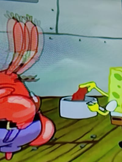 The image depicts a cartoon scene with two characters. In the foreground, on the left, is a large, red, amorphous character with purple pants and a black belt, possibly Mr. Krabs from SpongeBob SquarePants. Its right arm is raised, and its hand is blurred, suggesting rapid motion. Behind this character, on the right, is a yellow, porous character with green polka dots, likely SpongeBob SquarePants. SpongeBob is holding a red object with its right hand and appears to be dipping it into a white bowl with its left hand. The background is a light blue wall with some dark speckles. The floor is made of wooden planks, and there are some dark, round objects on the floor. The scene appears to be indoors, possibly in a kitchen or workshop setting. There is no discernible information within the image to indicate its location in Merbopuntong, Indonesia.