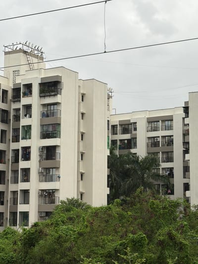 The image captures an overcast sky above a cluster of modern, multi-story residential buildings in Vasai-Virar, India. The buildings, primarily white and grey, feature numerous balconies with black railings and large windows. Some balconies display potted plants and laundry hanging to dry. Air conditioning units are visible on some exterior walls. A dense canopy of green trees and foliage fills the foreground, partially obscuring the lower levels of the buildings. In the background, a network of power lines crisscrosses the sky. One building's rooftop is equipped with numerous satellite dishes mounted on a metal frame, suggesting a significant number of residents utilizing satellite television services. The overall scene depicts a typical urban residential environment in a densely populated area.