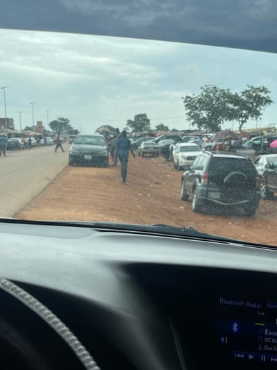 The image appears to have been taken from inside a vehicle in Karshi, Nigeria. It shows a dirt road crowded with parked cars and pedestrian traffic. A tree is visible in the background. A display screen of a car infotainment system is visible with “Bluetooth Audio” and audio track information.